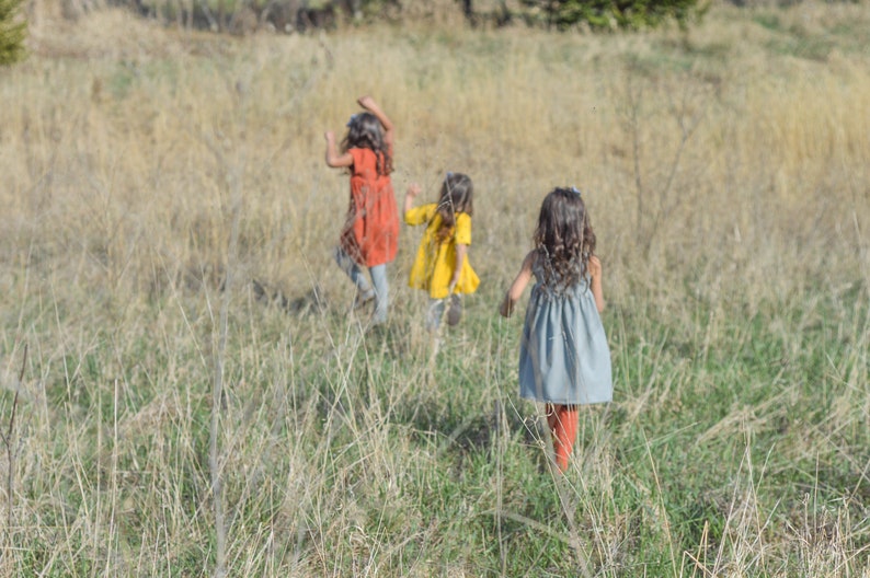 May include: Three young girls in dresses run through a field of tall grass. The girl in the front is wearing a blue dress, the middle girl is wearing a yellow dress, and the girl in the back is wearing an orange dress.
