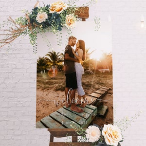 May include: A welcome sign with a photo of a couple embracing on a wooden dock. The sign is decorated with flowers and the text "welcome adam & jessica june 2021".
