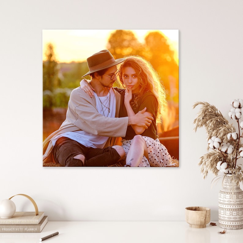 Op de afbeelding: Een verliefd stel, een man met een bruine hoed en een vrouw met lang blond haar, zitten samen in een veld bij zonsondergang. De man heeft zijn arm om de vrouw heen en ze kijken elkaar aan.