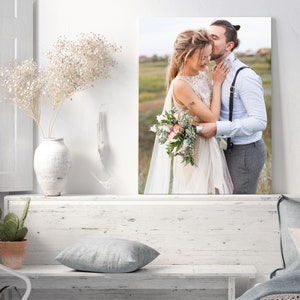 Op de afbeelding: Een trouwfoto van een stel dat elkaar omhelst in een veld. De bruid draagt een witte jurk en houdt een boeket bloemen vast. De bruidegom draagt een lichtblauw overhemd en een grijze broek.