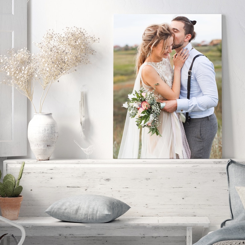 Op de afbeelding: Een trouwfoto van een stel dat elkaar omhelst in een veld. De bruid draagt een witte jurk en houdt een boeket bloemen vast. De bruidegom draagt een lichtblauw overhemd en een grijze broek.
