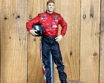 May include: A red racing suit figurine with a black helmet, standing on a silver beer tap. The figurine is wearing a red racing suit with black pants and white accents. The suit has various logos and sponsors printed on it.
