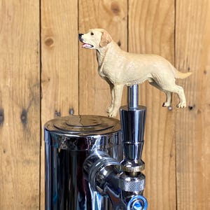 May include: A chrome beer tap with a detailed, tan-colored Labrador Retriever dog figurine on top. The tap has a shiny, reflective surface and is set against a wooden background.