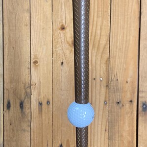 May include: A golf club with a brown grip and a light blue golf ball. The club is positioned vertically against a wooden background. The golf ball is positioned in the middle of the club.