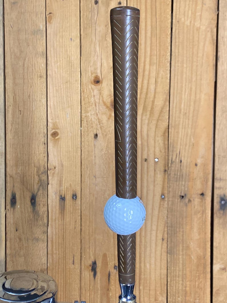 May include: A golf club with a brown grip and a light blue golf ball. The club is positioned vertically against a wooden background. The golf ball is positioned in the middle of the club.