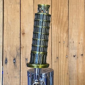 May include: A beer tap handle shaped like the Leaning Tower of Pisa. The handle is bronze and gold in color, with intricate architectural details. The tap itself is chrome, with a polished, reflective surface. The background is a wooden wall.