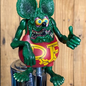 May include: A green cartoon character with large ears, bulging eyes, and sharp teeth. The figure wears a red top with yellow lettering and gives a thumbs-up. It is mounted on a chrome tap handle, likely for a keg.
