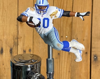 May include: A decorative tap handle featuring a football player in a white and blue uniform, number 20, in mid-air. The figure is mounted on a chrome tap, set against a wooden background. The player is holding a football.