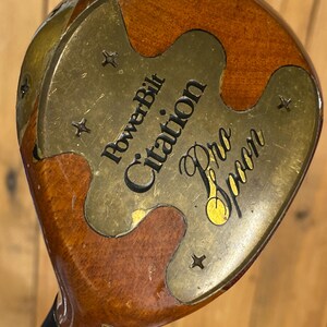 Puede incluir: Un palo de golf vintage PowerBilt Citation Pro Spoon. La cabeza del palo es una combinaci&oacute;n de madera y lat&oacute;n, con el texto "PowerBilt Citation Pro Spoon" grabado en la placa de lat&oacute;n. La madera tiene un acabado marr&oacute;n oscuro.