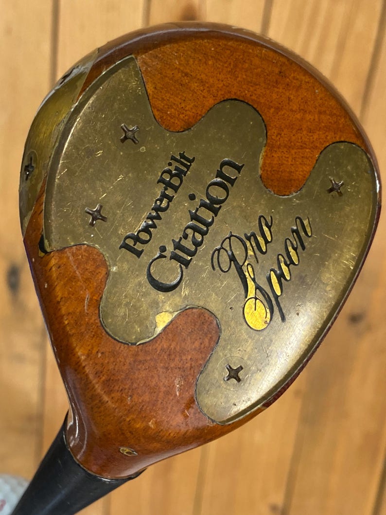 Puede incluir: Un palo de golf vintage PowerBilt Citation Pro Spoon. La cabeza del palo es una combinaci&oacute;n de madera y lat&oacute;n, con el texto "PowerBilt Citation Pro Spoon" grabado en la placa de lat&oacute;n. La madera tiene un acabado marr&oacute;n oscuro.