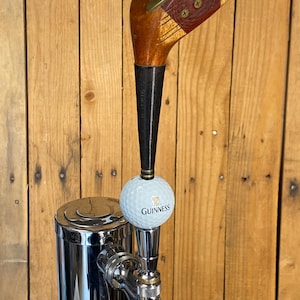Puede incluir: Un tirador de grifo de cerveza personalizado con una cabeza de palo de golf vintage y un mango de madera oscura. Se adjunta una pelota de golf con el logotipo de Guinness. El grifo es de metal pulido y est&aacute; montado sobre una superficie de madera.