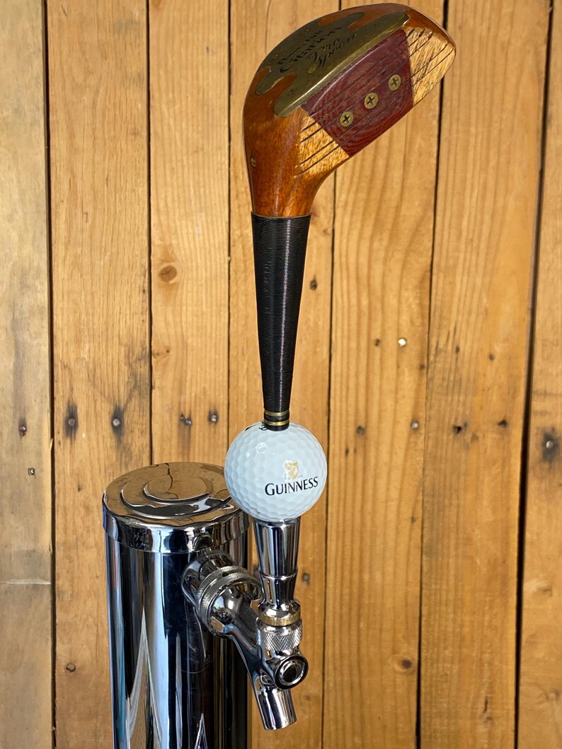 Puede incluir: Un tirador de grifo de cerveza personalizado con una cabeza de palo de golf vintage y un mango de madera oscura. Se adjunta una pelota de golf con el logotipo de Guinness. El grifo es de metal pulido y est&aacute; montado sobre una superficie de madera.