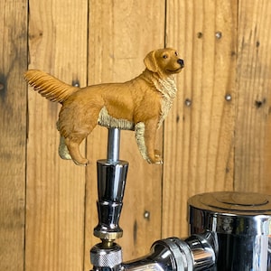 May include: A golden retriever dog figurine on top of a chrome beer tap. The tap is attached to a chrome beer dispenser.