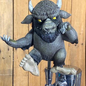 May include: A Where the Wild Things Are-themed tap handle featuring a Max character figurine. The figurine is gray and brown with yellow eyes, white horns, and a detailed fur texture. The tap handle is made of chrome and has a wooden base.