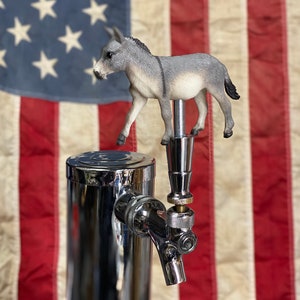 May include: A silver beer tap with a grey donkey figurine on top. The tap is in front of an American flag.