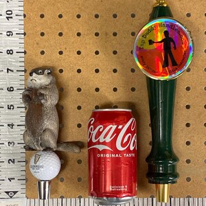 May include: An assortment of items including a groundhog figurine, a Guinness golf ball bottle stopper, a Coca-Cola can, and a green tap handle with the text "Tap Daddy Handles.com". The Coca-Cola can is red with white text.
