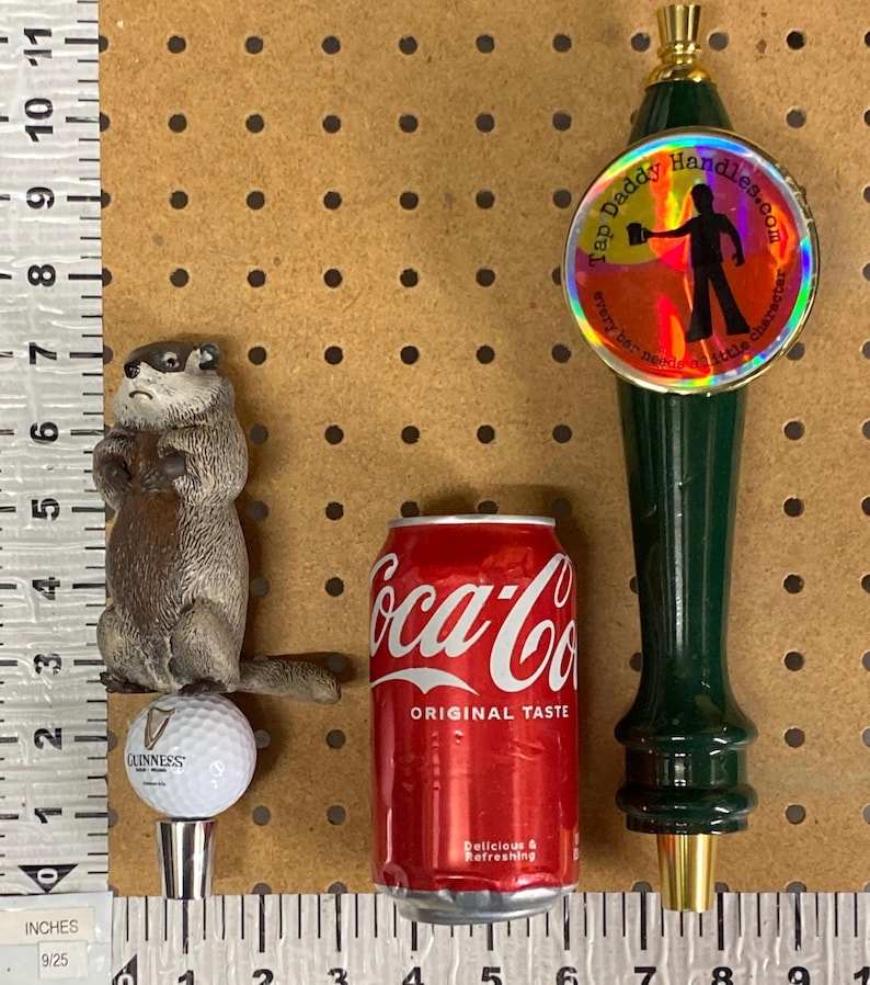 May include: An assortment of items including a groundhog figurine, a Guinness golf ball bottle stopper, a Coca-Cola can, and a green tap handle with the text "Tap Daddy Handles.com". The Coca-Cola can is red with white text.