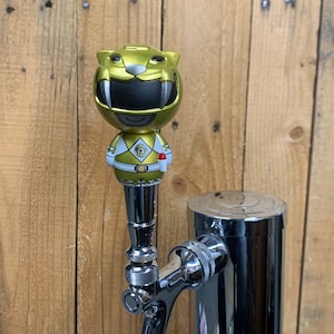 May include: Gold Power Rangers character tap handle with a yellow and white suit and a black helmet. The character is a tiger. The tap handle is attached to a chrome beer tap.