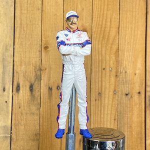 May include: A white and blue racing driver figurine with a black mustache and sunglasses, wearing a white cap with a red, white, and blue logo. The figurine is mounted on a silver beer tap.