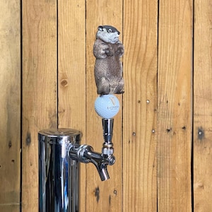 May include: A chrome beer tap with a groundhog tap handle. The groundhog is brown and gray, standing on a golf ball with the Guinness logo. The tap is set against a wooden background.