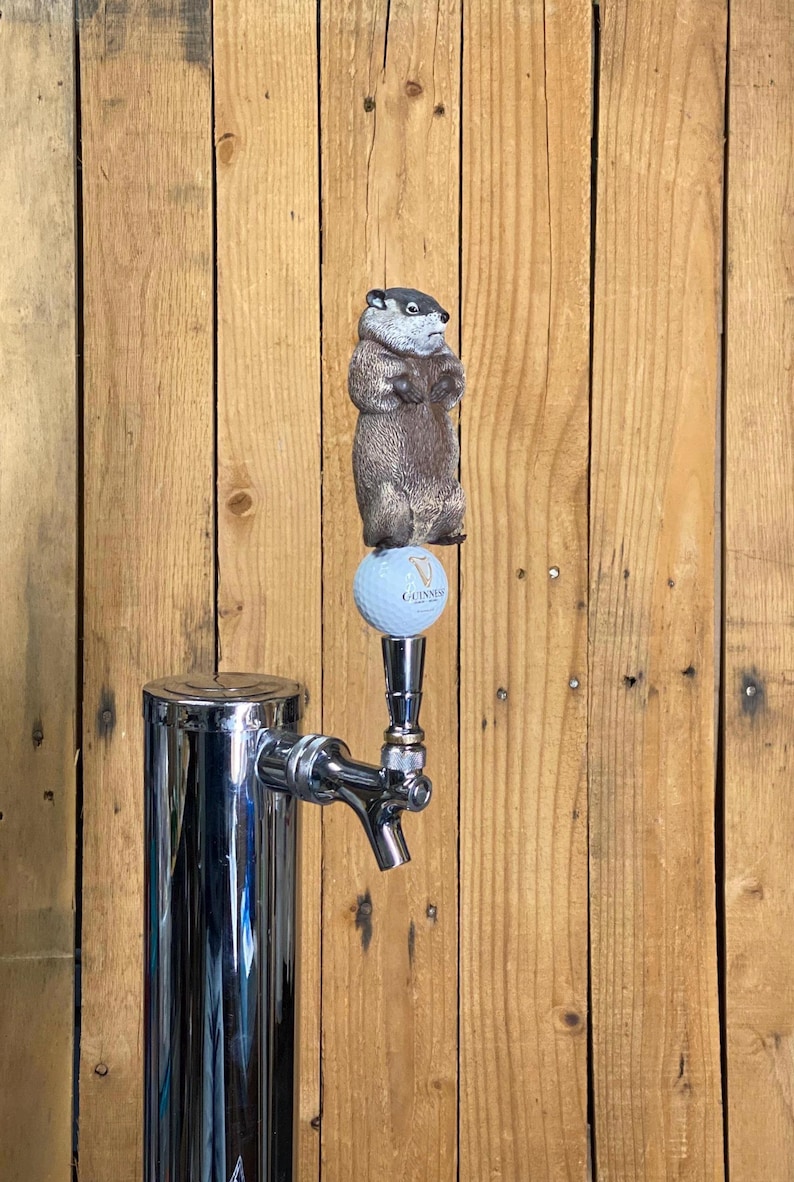 May include: A chrome beer tap with a groundhog tap handle. The groundhog is brown and gray, standing on a golf ball with the Guinness logo. The tap is set against a wooden background.