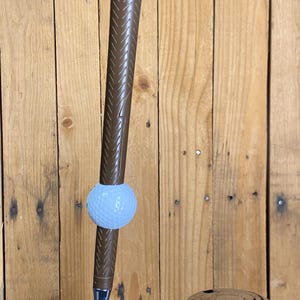 May include: A brown golf club tap handle with a golf ball attached. The handle has a textured grip and is connected to a chrome tap. The background is a wooden wall.