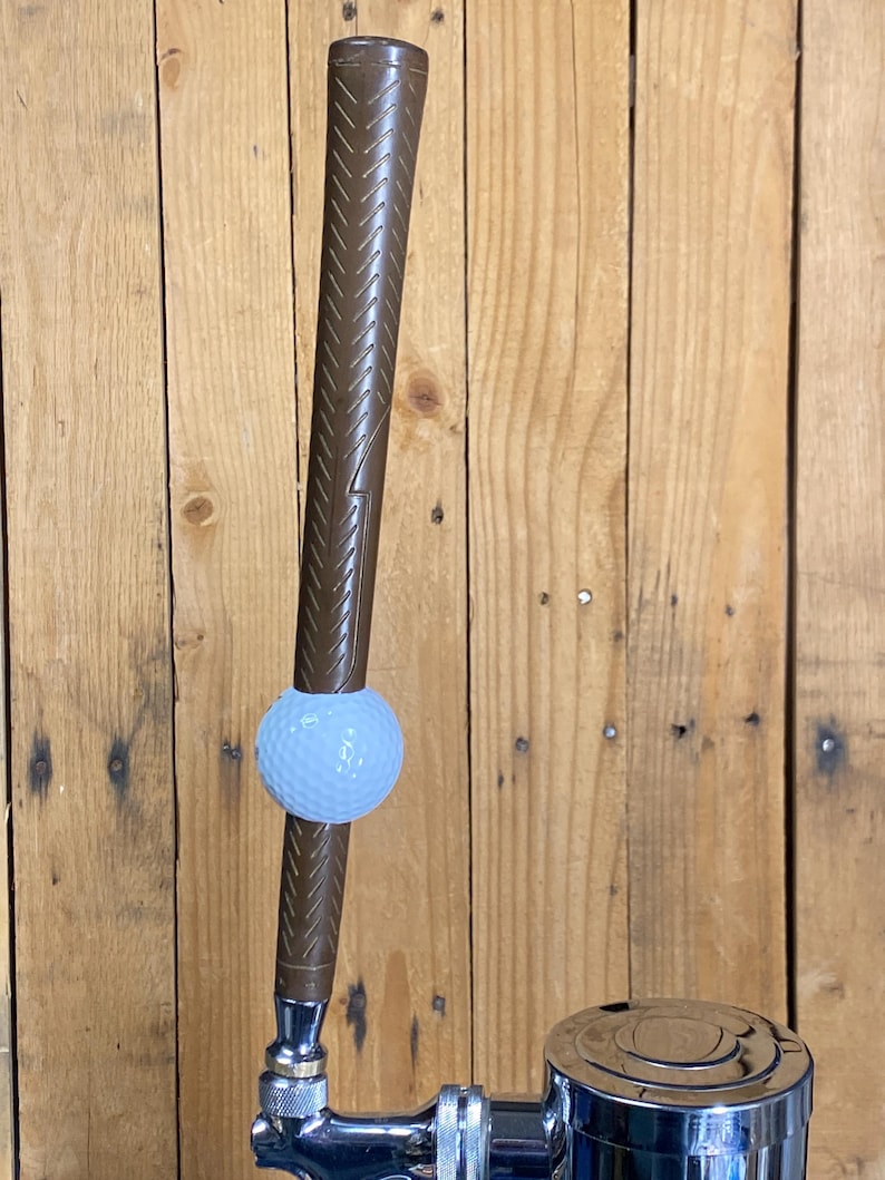 May include: A brown golf club tap handle with a golf ball attached. The handle has a textured grip and is connected to a chrome tap. The background is a wooden wall.