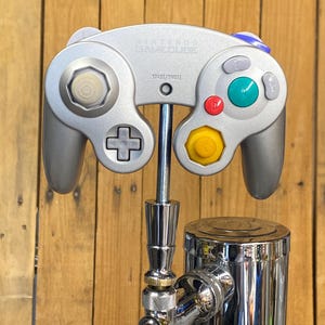 May include: A silver Nintendo GameCube controller mounted on a chrome beer tap. The controller has a gray body with colorful buttons and the text "Nintendo GameCube". The tap is set against a wooden background.