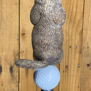 May include: A golf club accessory featuring a detailed, brown and tan groundhog figurine perched atop a light blue golf ball. The groundhog has a textured fur appearance. The golf ball sits on a silver-colored metal shaft.