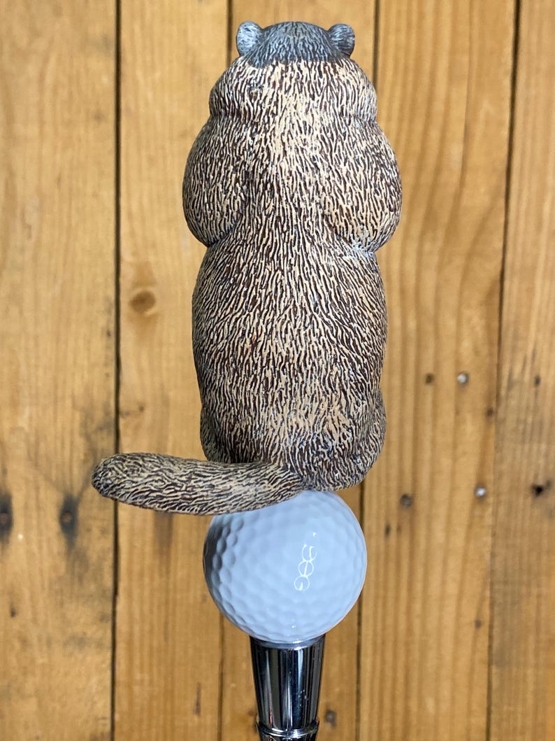May include: A golf club accessory featuring a detailed, brown and tan groundhog figurine perched atop a light blue golf ball. The groundhog has a textured fur appearance. The golf ball sits on a silver-colored metal shaft.