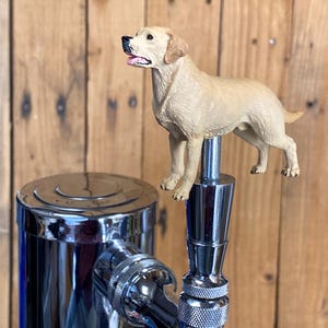 May include: A beer tap handle featuring a sculpted, light tan Labrador retriever dog figure standing atop a polished chrome metal base. The dog has a black nose, open mouth with a visible pink tongue, and textured fur detail throughout its body.