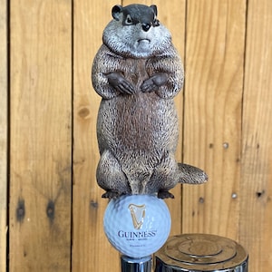 May include: A groundhog-themed beer tap handle. The groundhog is brown and gray, standing on a golf ball with the Guinness logo. The tap handle is made of metal and has a shiny, silver finish. The background is a wooden wall.