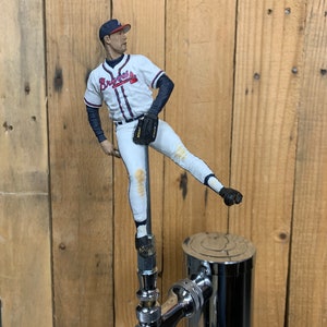 May include: A baseball player figurine wearing a white and red baseball uniform with the word "Braves" on the front, holding a baseball glove, mounted on a chrome beer tap.