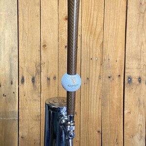May include: A chrome beer tap with a brown handle and a golf ball-shaped accent featuring the Guinness logo. The tap is set against a wooden background.