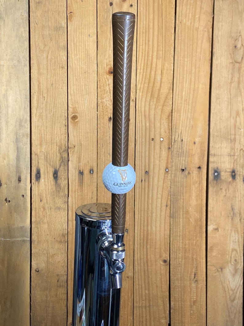 May include: A chrome beer tap with a brown handle and a golf ball-shaped accent featuring the Guinness logo. The tap is set against a wooden background.