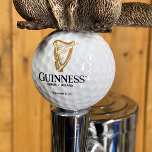 May include: A beer tap handle featuring a brown beaver figure atop a white golf ball with the Guinness logo. The tap handle has a silver metal base and a wooden background.