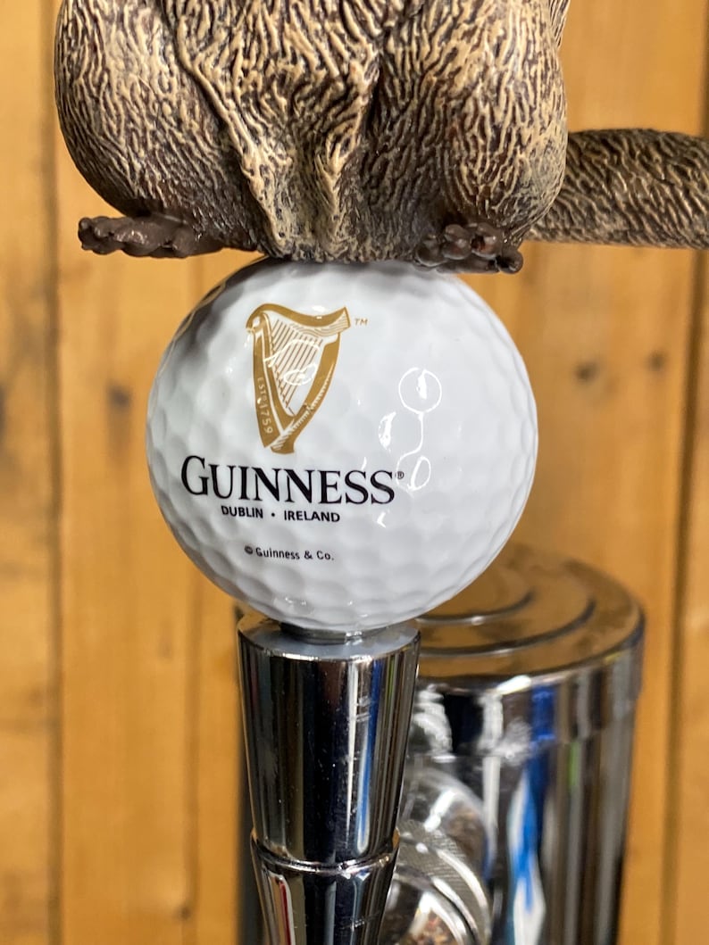 May include: A beer tap handle featuring a brown beaver figure atop a white golf ball with the Guinness logo. The tap handle has a silver metal base and a wooden background.