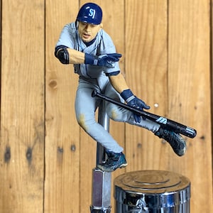 May include: A baseball-themed tap handle featuring a figurine of a baseball player in a gray uniform and blue cap. The player is in mid-swing, holding a black bat. The tap handle has a chrome finish and is mounted on a wooden background.