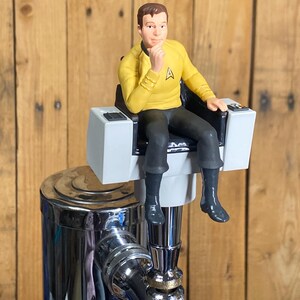 May include: A yellow and black Star Trek-themed tap handle featuring a figure in a command chair. The figure is wearing a yellow uniform with a black collar and black pants. The tap handle is mounted on a chrome tap.