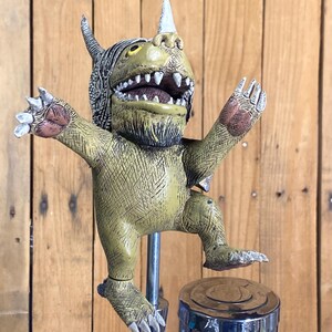 May include: A whimsical, olive green tap handle featuring a monster character with a white horn, yellow eyes, and open mouth. The monster has outstretched arms and detailed claws. The handle is mounted on a chrome tap.