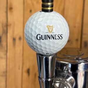 Puede incluir: Un tirador de grifo blanco con forma de pelota de golf con el logotipo de Guinness y un arpa. El tirador tiene una parte superior negra y dorada y una base de metal brillante, sobre un fondo de madera.