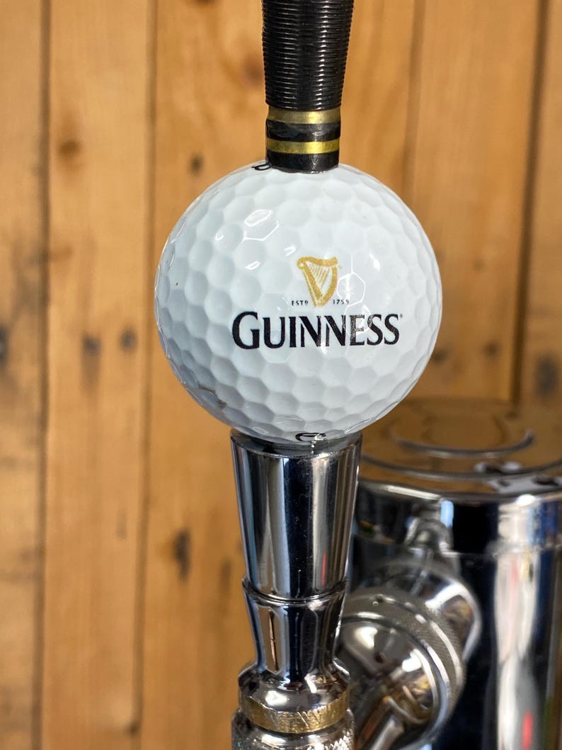 Puede incluir: Un tirador de grifo blanco con forma de pelota de golf con el logotipo de Guinness y un arpa. El tirador tiene una parte superior negra y dorada y una base de metal brillante, sobre un fondo de madera.
