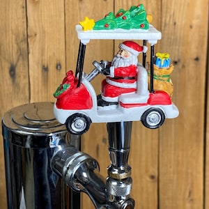 May include: A Christmas-themed tap handle featuring Santa Claus driving a red and white golf cart. The cart has a green Christmas tree on top and a bag of gifts in the back. The tap handle is attached to a chrome beer tap.