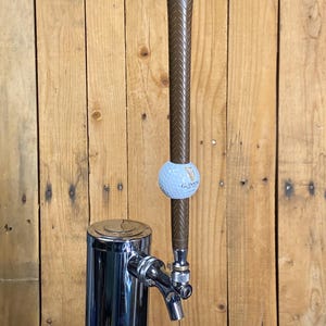 May include: A chrome beer tap with a brown golf club handle and a golf ball with the Guinness logo. The tap is mounted on a wooden background.