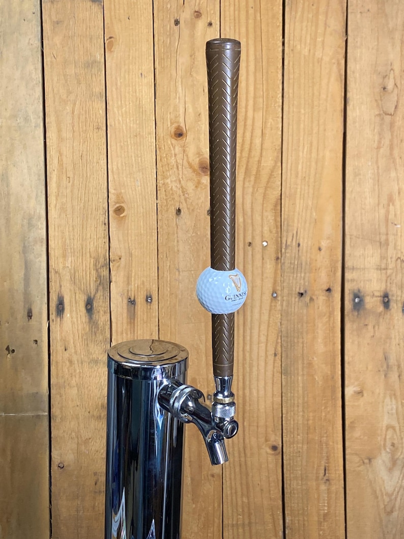 May include: A chrome beer tap with a brown golf club handle and a golf ball with the Guinness logo. The tap is mounted on a wooden background.