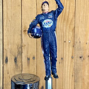 May include: A blue and white figurine of a race car driver in a racing suit, holding a helmet. The figurine is mounted on a chrome tap handle. The driver is waving with his right hand.