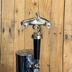 May include: A silver-colored beer tap handle with a small, detailed sculpture of a race car driver in a vintage race car. The handle is black and has a chrome base.