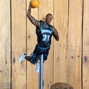 May include: A figurine of a basketball player in a black and teal jersey with the number 21, holding a basketball. The figure is mounted on a metal pole, with a wooden background.