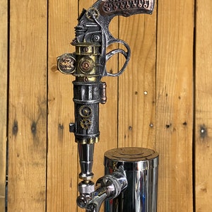 May include: A steampunk-style beer tap handle in the shape of a pistol with intricate details and a copper-colored grip. The tap handle is attached to a chrome beer tap.