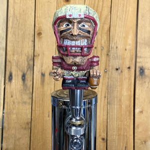 May include: A wooden football player tap handle with a smiling face, wearing a maroon and tan helmet and jersey. The handle is mounted on a shiny, chrome-colored tap.